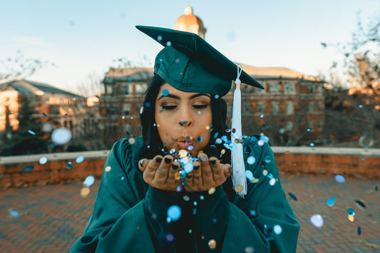 A woman wearing a graduation cap and gown blows confetti toward the camera. Photo by Clay Banks for Unsplash